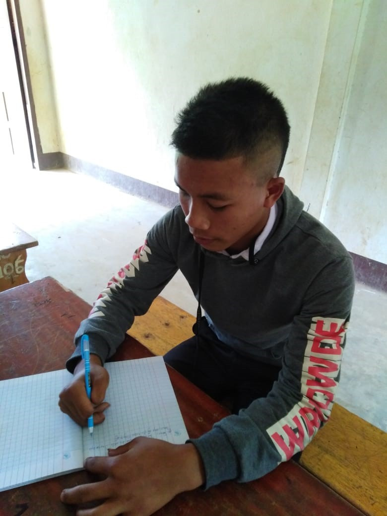 Ein junger Mann sitzt an einem Tisch und schreibt etwas in ein Heft. An der linken Hand sind einige Finger angeschnitten.