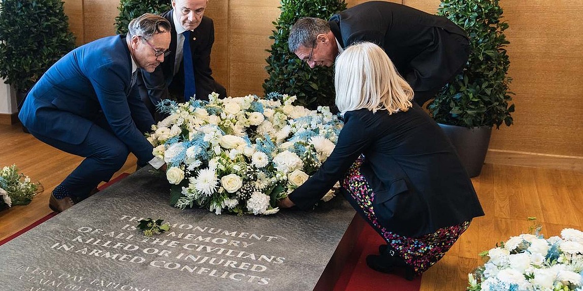 Drei Männer und eine Frau legen auf einem Grabstein mit Inschrift einen Blumenkranz nieder.