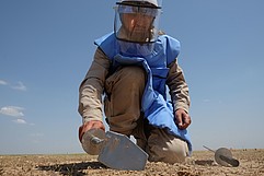 Ein Minenräumer kniet in Schutzkleidung und mit Werkzeug auf sandigem Boden.