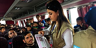 Eine Frau steht in einem vollen Bus und verteilt Flyer auf denen Minenaufklärung zu sehen ist.