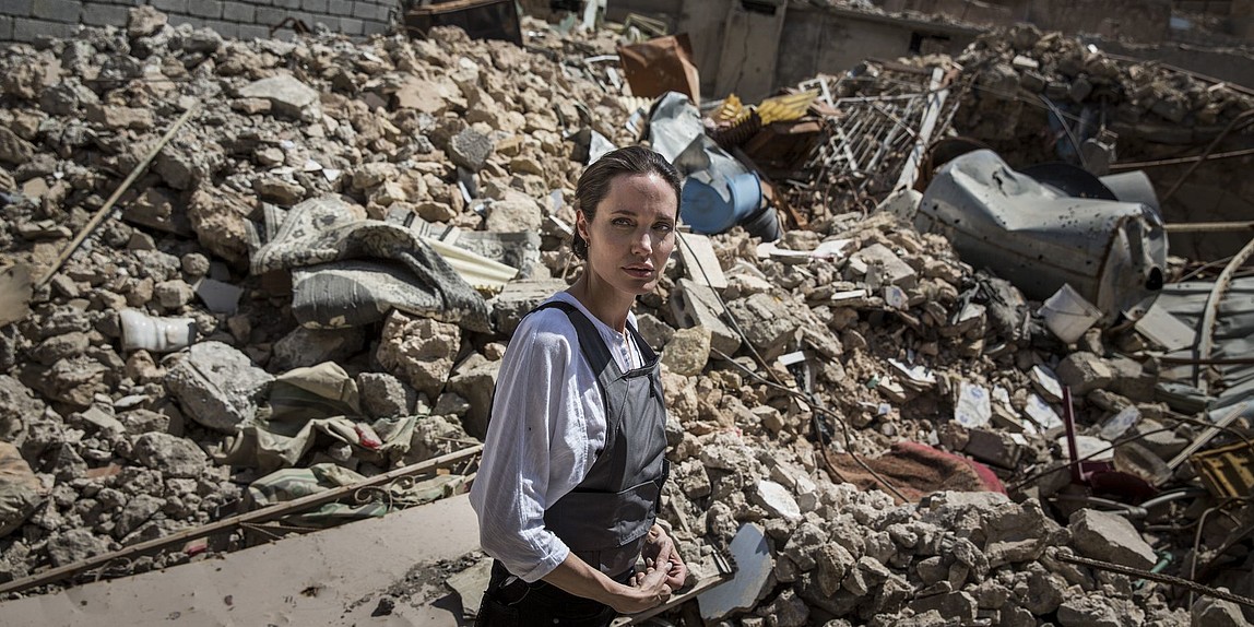 Eine Frau in Hemd und Minenschutzschürze steht inmitten von Trümmern und blickt sich besorgt nach hinten um. 
