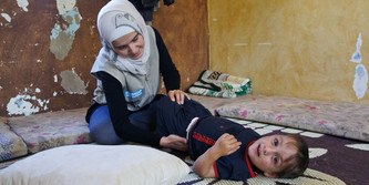 Eine Mitarbeiterin von Handicap International kniet auf dem Boden und macht Übungen mit Fayez. Dieser liegt auf dem Rücken und lacht in die Kamera.