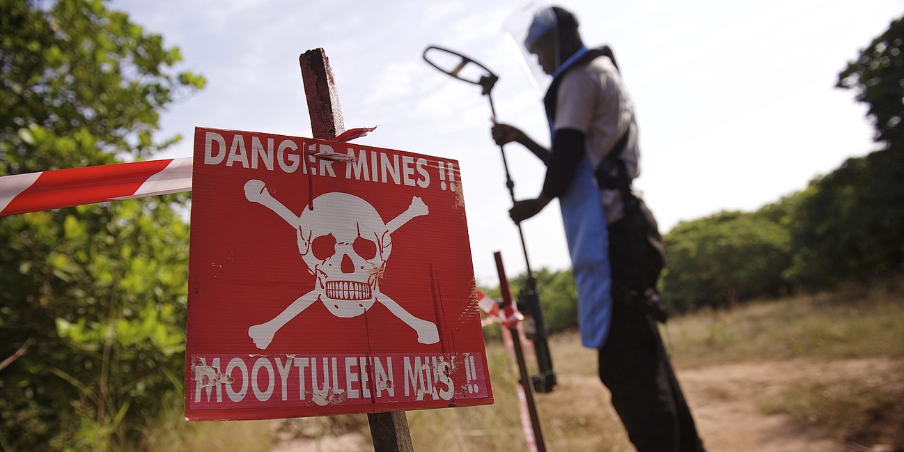 Im Vordergrund ein Danger Mines Schild, im Hintergrund ein Mann mit Entminerausrüstung