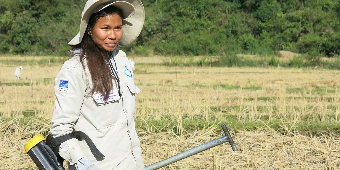 Eine Frau mit Arbeitskleidung steht mit einem Minendetektor in einem Feld.