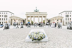 Vorne liegt das Mahnmal mit einem Kranz. Dahinter stehen Menschen in einem Halbkreis und halten sehr große Portraits hoch. Im Hintergrund das Brandenburger Tor.