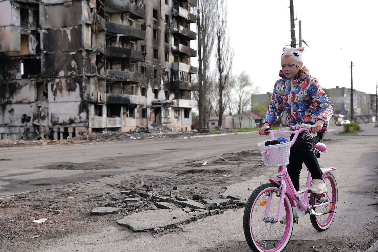 Ein Mädchen fährt auf einem Fahrrad vor einem völlig zerstörten Hochhaus.